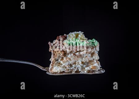 Fetta di torta con torte di tipo meringa con noci proteiche con uno strato e decorazione di crema crema crema di burro su spatola metallica su sfondo nero, primo piano Foto Stock