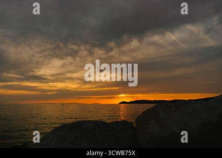 Un tramonto mozzafiato sul mare, con il sole che si immerge dietro un'isola o una penisola dalle linee scure. Il primo piano presenta grandi rocce scure Foto Stock