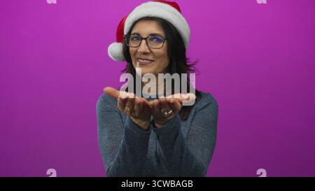 Una donna ispanica di mezza età sorridente tiene le palme aperte rivolte verso l'alto e indossa il cappello di babbo natale in studio; generosità per le vacanze. Foto Stock