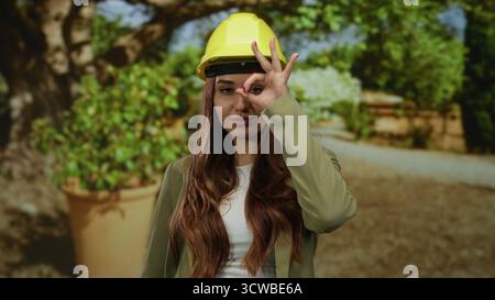 Una donna che indossa un elmetto fa un buon gesto nel parco soleggiato, mescolando lavoro e svago come architetto o ingegnere in un ambiente all'aperto vivace, incarnando pr Foto Stock
