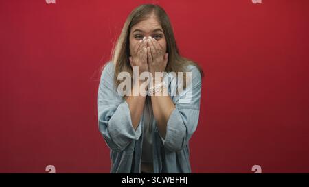 La giovane donna bionda alza le mani in studio davanti a uno sfondo rosso che mostra un gesto di bocca aperta con gli occhi larghi; stupore. Foto Stock