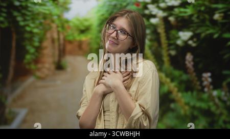 Una donna bionda che indossa gli occhiali tiene delicatamente le mani sul petto sotto il baldacchino frondoso nella foresta verde; gratitudine. Foto Stock