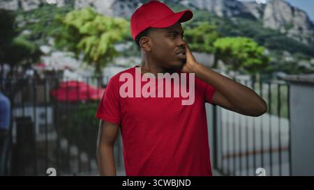 Uomo afroamericano che indossa un berretto rosso e una camicia che preme la mano contro l'orecchio per l'orecchio sulla strada urbana accanto alla ringhiera di metallo; disagio. Foto Stock