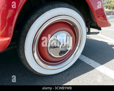 Primo piano di una ruota d'auto d'epoca con pneumatico whitewall e coprimozzo cromato con centro rosso, parcheggiata su asfalto con linee di parcheggio visibili. Foto Stock