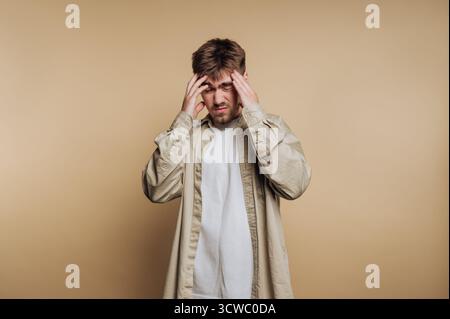 il ritratto di un giovane uomo esprime frustrazione tenendo la testa, catturando un momento emotivo su sfondo beige Foto Stock