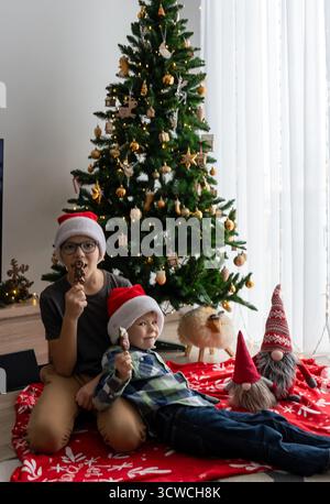 Due fratelli nei cappelli di Babbo Natale che mangiano biscotti al cioccolato di pan di zenzero seduti vicino all'albero di Natale decorato con ornamenti dorati e un giocattolo natalizio. Totale Foto Stock
