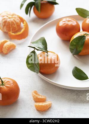 Mandarini vivaci con foglie verdi, che mostrano freschezza e fascino agrumato su un piatto bianco e un tavolo Foto Stock