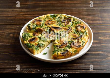 Frittata con spinaci e funghi champignon su piatto bianco su fondo marrone in legno. Vista ravvicinata Foto Stock