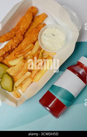 Una porzione di patatine fritte e strisce di pollo con salsa in una scatola di cartone da asporto. Bastoncini di pollo croccanti e patate fritte con ketchup su anatra blu Foto Stock