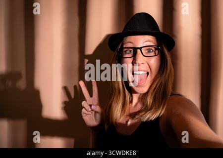 Una giovane donna che indossa un cappello e occhiali sta facendo un segno di pace mentre si fa un selfie Foto Stock