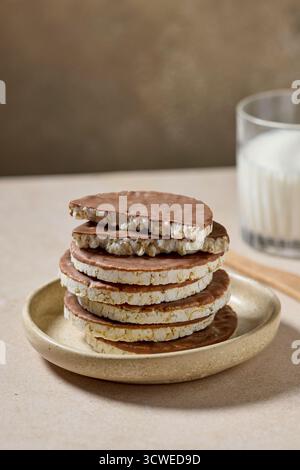 una pila di crocchette di riso torte e biscotti ricoperti di cioccolato al latte e un bicchiere di latte su sfondo beige Foto Stock