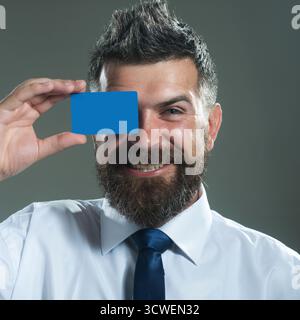 Un uomo d'affari sorridente che copre l'occhio con carta di credito o di debito. E-commerce, pagamenti senza contanti. Finanza, e-money. Servizi bancari. Uomo barbuto di successo Foto Stock