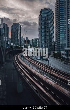 Gli alti edifici delimitano la città mentre i binari del treno si snodano attraverso le strade trafficate durante il crepuscolo, catturando la vita urbana al crepuscolo. Foto Stock
