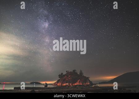 Una tranquilla scena costiera di notte mostra una piccola isola illuminata da stelle e un debole bagliore, circondata da acque calme e luci lontane. Foto Stock