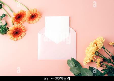Busta con carta bianca, fiori di gerbera arancio e crisantemi gialli su sfondo rosa corallo. Mockup floreale per messaggio o invito. Vista dall'alto Foto Stock