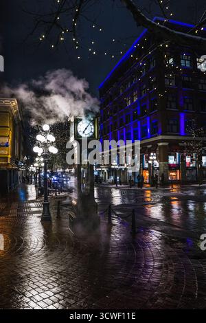 La scena mostra una torre dell'orologio d'epoca circondata da luci accese e riflessi sul marciapiede bagnato durante una serata di pioggia. Foto Stock
