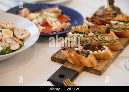 Foto ravvicinata di un piatto di bruschette disposto ad arte con vari condimenti su un tavolo bianco, che presenta rustiche tavole di legno e piatti blu, alti Foto Stock