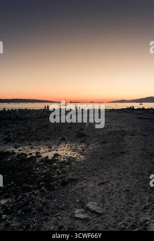 Le persone camminano lungo la riva mentre il sole tramonta sull'acqua, creando una colorata scena crepuscolare con sagome all'orizzonte. Foto Stock