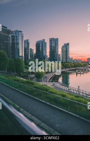 Gli edifici torreggianti riflettono il colorato tramonto di Vancouver, con una tranquilla passeggiata lungo l'acqua piena di camminatori serali. Foto Stock