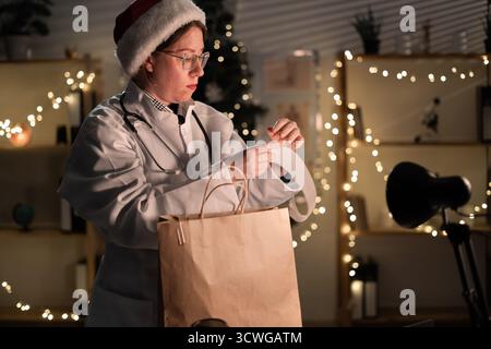Giovane medico donna in occhiali da vista e cappello di Babbo Natale durante il turno disimballaggio cibo da asporto in sacchetto di carta lavoro di notte la vigilia di Natale in ospedale. Foto Stock