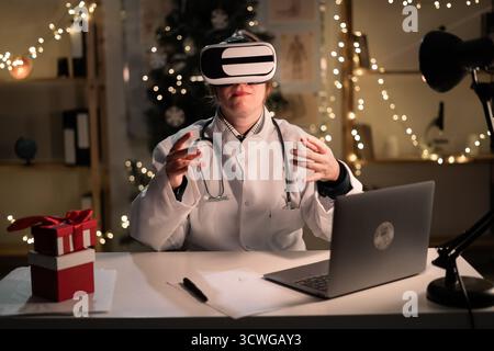 La giovane dottoressa indossa il cappello di Babbo Natale e gli occhiali vr che guardano la realtà metaversa durante il turno di lavoro la sera della vigilia di Natale. Medicina, santo Foto Stock