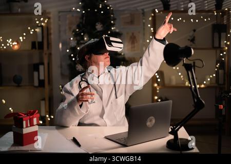 La giovane dottoressa indossa il cappello di Babbo Natale e gli occhiali vr che guardano la realtà metaversa durante il turno di lavoro la sera della vigilia di Natale. Medicina, vacanze, Foto Stock