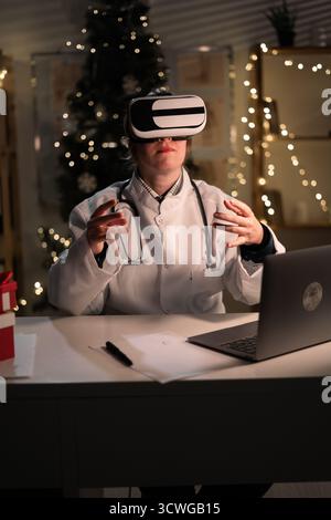 La giovane dottoressa indossa il cappello di Babbo Natale e gli occhiali vr che guardano la realtà metaversa durante il turno di lavoro la sera della vigilia di Natale. Medicina, santo Foto Stock