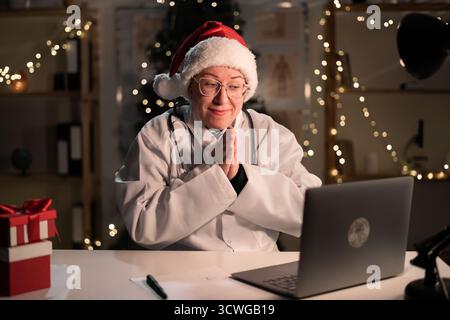 La giovane dottoressa indossa il cappello di Babbo Natale e gli occhiali da vista usando un computer portatile che tiene le mani in mano aiutami, per favore, gesto durante il turno di notte su Cristo Foto Stock