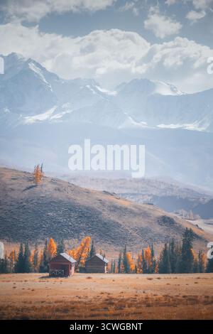 In una valle tranquilla, due capanne rustiche sorgono circondate da vibranti alberi autunnali e colline ondulate, sotto uno sfondo di maestose montagne. Nuvole Foto Stock