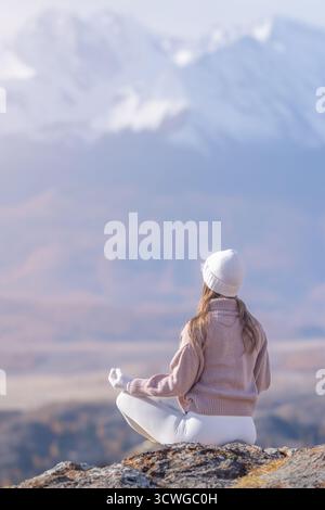 Una donna vestita calorosamente siede su una roccia, meditando pacificamente sulle montagne autunnali. Gode del tranquillo paesaggio circondato da maestose cime e. Foto Stock