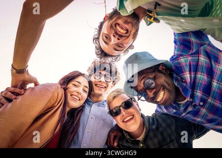 Il punto di vista ad angolo basso di un gruppo felice di amici giovani e diversi, rannicchiati in cerchio e sorridenti davanti alla telecamera all'aperto. Foto Stock