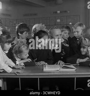 Il celebre capitano rompighiaccio sovietico Anatoly Lamekhov, eroe dell'Unione Sovietica, è circondato da affascinanti studenti delle scuole elementari in questa emozionante foto degli anni '1980. Il famoso esploratore artico, indossando la sua uniforme con la medaglia Gold Star, condivide le storie di un libro aperto durante una lezione di educazione patriottica nella sua città natale, Sloviansk, RSS Ucraina. I bambini, nelle loro uniformi scolastiche, ascoltano con ammirazione. Questa immagine cattura una tipica "lezione di coraggio" e l'ispirazione di un eroe che incontra la prossima generazione in URSS Foto Stock