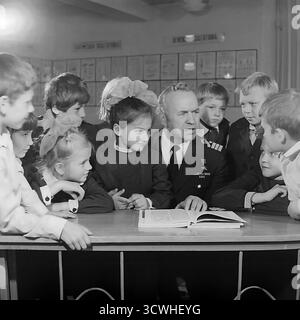 Il celebre capitano rompighiaccio sovietico Anatoly Lamekhov, eroe dell'Unione Sovietica, è circondato da affascinanti studenti delle scuole elementari in questa emozionante foto degli anni '1980. Il famoso esploratore artico, indossando la sua uniforme con la medaglia Gold Star, condivide le storie di un libro aperto durante una lezione di educazione patriottica nella sua città natale, Sloviansk, RSS Ucraina. I bambini, nelle loro uniformi scolastiche, ascoltano con ammirazione. Questa immagine cattura una tipica "lezione di coraggio" e l'ispirazione di un eroe che incontra la prossima generazione in URSS Foto Stock