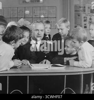 Il celebre capitano rompighiaccio sovietico Anatoly Lamekhov, eroe dell'Unione Sovietica, è circondato da affascinanti studenti delle scuole elementari in questa emozionante foto degli anni '1980. Il famoso esploratore artico, indossando la sua uniforme con la medaglia Gold Star, condivide le storie di un libro aperto durante una lezione di educazione patriottica nella sua città natale, Sloviansk, RSS Ucraina. I bambini, nelle loro uniformi scolastiche, ascoltano con ammirazione. Questa immagine cattura una tipica "lezione di coraggio" e l'ispirazione di un eroe che incontra la prossima generazione in URSS Foto Stock