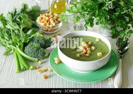 Purea di zuppa sana di broccoli, sedano ed erbe con crostini in ciotola bianca pronti da mangiare accanto agli ingredienti su sfondo di legno bianco. Primo piano. Foto Stock
