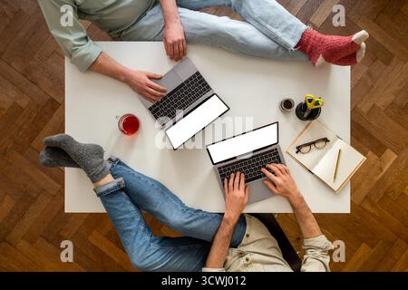 Vista dall'alto di due colleghi nello Smart Working. Notebook con schermo bianco per il mockup. Piedi sulla scrivania e notebook sul tavolo. Collaborazione Foto Stock