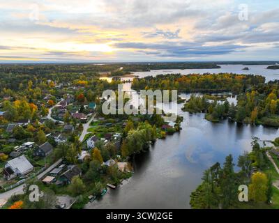 Vista aerea di un fiume tortuoso che passa attraverso un piccolo villaggio e una fitta foresta con un vivace fogliame autunnale che conduce a un grande lago Foto Stock