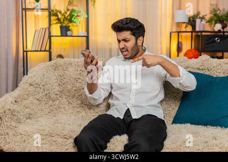 L'uomo indiano a casa sul divano afferrando lo smartphone ermeticamente, sconvolto, con rabbia buttandolo da parte dopo cattive notizie. Ragazzo arabo frustrato, infastidito, reagito emotivamente, mostrando stress, delusione, irritazione Foto Stock
