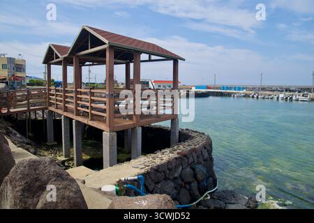 Un padiglione in legno sul lungomare su palafitte si affaccia sul porto di Beollang, con vasche di stoccaggio blu e barche ormeggiate che costeggiano il porto sotto nuvole sparse. Foto Stock