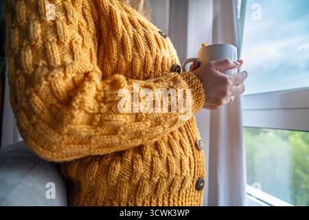 Persona che indossa un cardigan giallo mostarda e tiene una bevanda calda vicino alla finestra Foto Stock