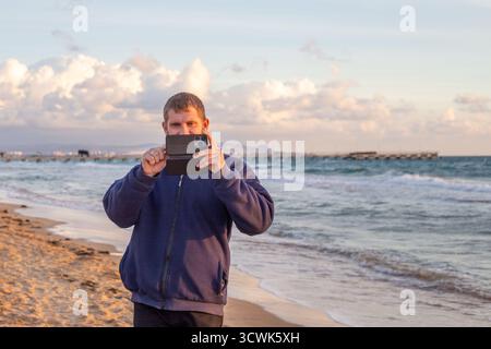 Un uomo si trova su una spiaggia sabbiosa, scattando una foto di una pittoresca vista sull'oceano sul suo telefono sotto un cielo nuvoloso e luminoso. Foto Stock