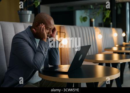 Uomo d'affari maturo e stressato con mal di testa che sfrega le stanghette con un laptop alla scrivania in ufficio Foto Stock