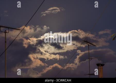 La luna lotta per rompere un denso gruppo di spettacolari nuvole notturne, incorniciate da una rete caotica di antenne sul tetto, fili e un camino in un Foto Stock