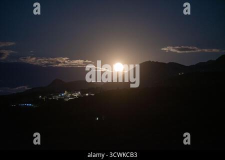 Una brillante luna piena sorge su una silhouette di montagna scura, che proietta un bagliore luminoso sul paesaggio e un piccolo villaggio greco illuminato sotto. A cl Foto Stock