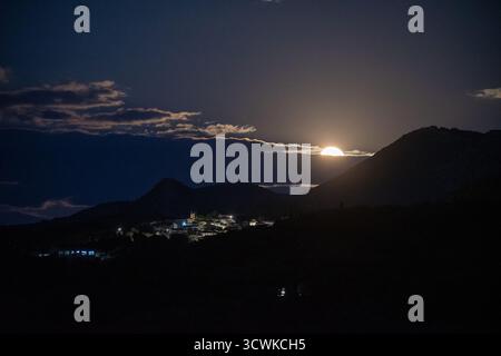 Spettacolare alba su una catena montuosa dalle linee sagomate, che rivela la luna piena sopra un luminoso villaggio greco. Un tranquillo e bellissimo paesaggio notturno si dispiega Foto Stock