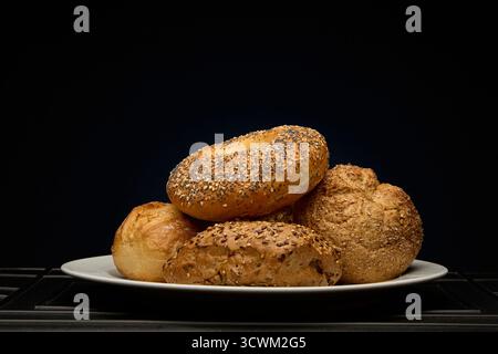 Panini freschi assortiti impilati su un piatto bianco su sfondo scuro illuminati da luce viola e ciano. Moderno concetto di panificio immagine con arte Foto Stock