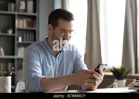 Giovane dipendente di ufficio allegro seduto alla scrivania che legge il telefono Foto Stock