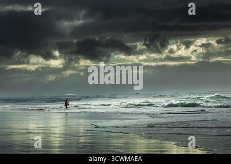 Un pescatore solitario si erge su una spiaggia, gettando una fila nel surf contro uno sfondo drammatico di cieli tempestosi. Le nuvole scure si stagliano in alto, a contrasto con Foto Stock