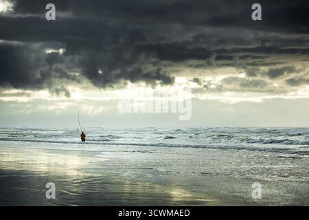 Un pescatore solitario si erge sul surf, gettando una fila nelle acque turbolente. La scena è drammatica, con un cielo buio e tempestoso in alto e nuvole pesanti Foto Stock