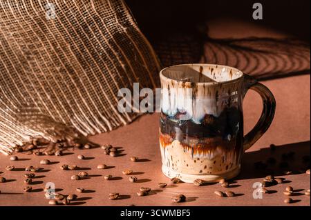 Una tazza da caffè in ceramica su sfondo marrone con chicchi di caffè in primo piano e luce del sole mattutino Foto Stock